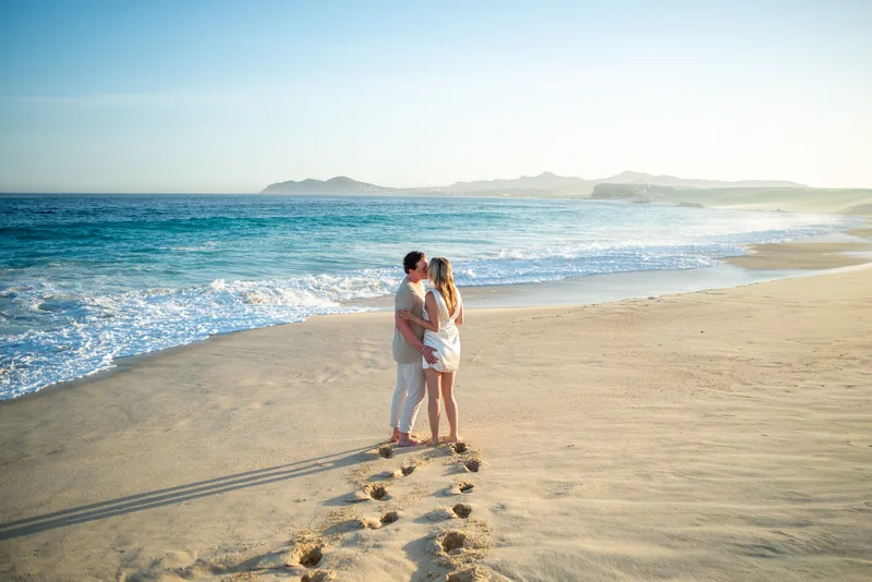 Jacquelyn and Jake kissing at the shoreline in Los Cabos, their footprints in the sand behind them